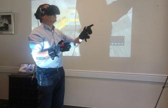 A white man in a corporate outfit with a headset and gloves using a virtual reality tool. in the background, we can see that he is playing in an industrial environment (on a computer screen far away and a projected image on the wall hidden by the man's shadow).